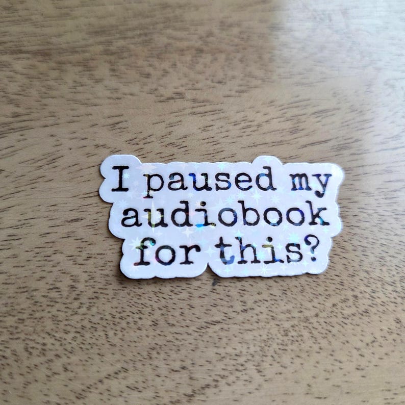 Pu&ograve; includere: Un adesivo fustellato con finitura olografica che riporta il testo nero "I paused my audiobook for this?". L'adesivo presenta un bordo bianco e un sottile motivo a stelle integrato nelle lettere su uno sfondo scintillante e riflettente.