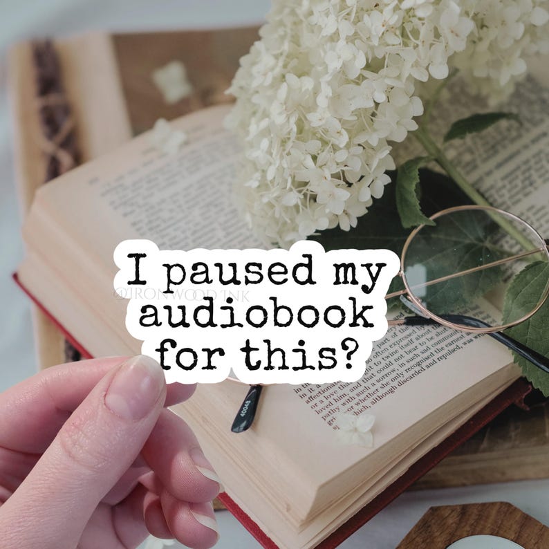 May include: A white die-cut sticker with black text that reads "I paused my audiobook for this?" positioned over an open book with cream-colored pages, a pair of thin-rimmed round glasses, and a cluster of white hydrangea flowers.