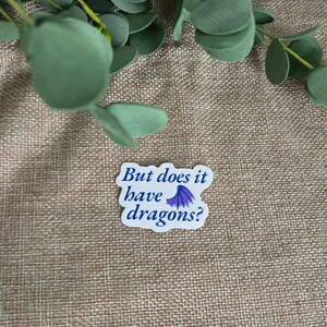 May include: A white die-cut sticker with a wavy edge featuring the dark blue text "But does it have dragons?" and a small purple dragon wing icon positioned between the words "have" and "dragons" on the final line of the printed message.