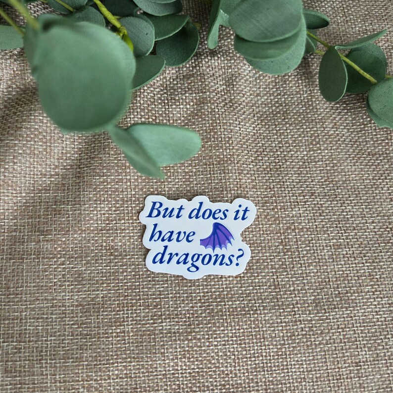 May include: A white die-cut sticker with a wavy edge featuring the dark blue text "But does it have dragons?" and a small purple dragon wing icon positioned between the words "have" and "dragons" on the final line of the printed message.