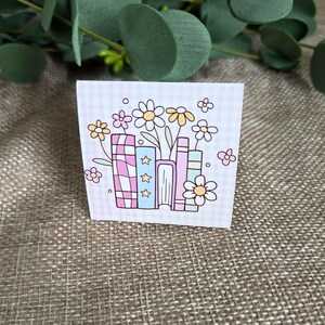 May include: A square sticker featuring a light pink gingham background with a central illustration of four books in pastel colors. The books are decorated with stars and stripes, surrounded by several yellow and pink daisies with green stems.