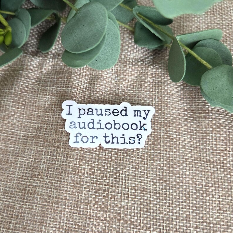 May include: A white die-cut sticker with a black typewriter-style font that reads, I paused my audiobook for this? The sticker features a slightly irregular, organic shape and is positioned against a textured, light brown fabric background.
