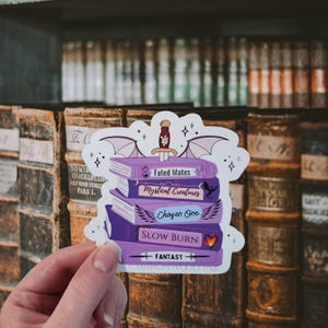 May include: A die-cut sticker featuring a stack of five purple books topped with a dagger and bat wings. The book spines are labeled Fated Mates, Mystical Creatures, Chosen One, SLOW BURN, and FANTASY, with small icons including a dragon and heart.