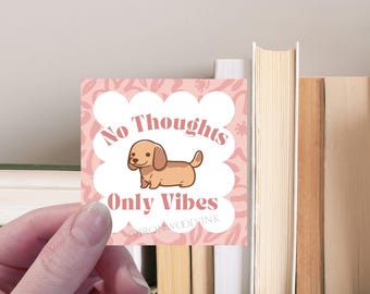 Può includere: Un adesivo quadrato con sfondo rosa chiaro caratterizzato da una forma smerlata bianca al centro. All'interno della forma c'è un bassotto marrone in stile cartone animato e il testo No Thoughts Only Vibes. Il testo IRONWOODINK è stampato in basso.