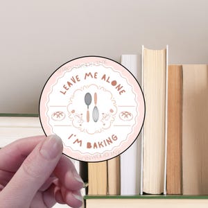 May include: Pink round sticker with the text "Leave me alone I'm baking" in brown lettering. The sticker has a white border and a design of two kitchen utensils and two loaves of bread.
