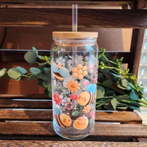 May include: A clear glass tumbler with a bamboo lid and a glass straw, featuring a colorful wrap-around print of cartoon bees, orange honeycomb patterns, small pink and orange flowers, and a small honey pot with the word Honey printed on it.