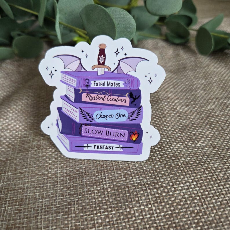 May include: A die-cut sticker featuring a stack of five purple books topped with a dagger and bat wings. The book spines are labeled Fated Mates, Mystical Creatures, Chosen One, Slow Burn, and Fantasy, decorated with small icons like a dragon and heart.