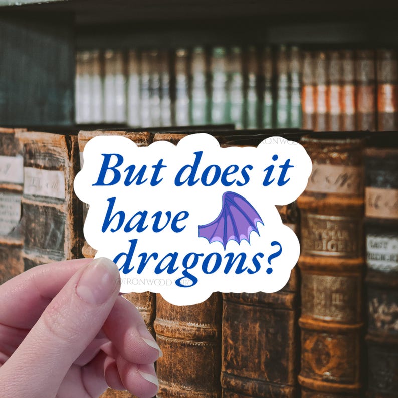 May include: A white die-cut sticker with a wavy edge featuring the blue text "But does it have dragons?" and a small purple dragon wing illustration. The sticker has a glossy finish and is held against a background of old leather-bound books.