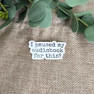 Pu&ograve; includere: Un adesivo bianco fustellato con un carattere nero in stile macchina da scrivere che recita: "I paused my audiobook for this?" L'adesivo ha una forma rettangolare arrotondata irregolare ed &egrave; posizionato su uno sfondo in tessuto marrone chiaro.