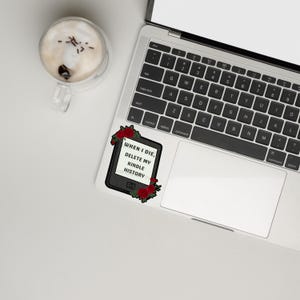 May include: A rectangular sticker featuring a black e-reader device surrounded by red roses and green leaves. The screen displays the black text: WHEN I DIE, DELETE MY KINDLE HISTORY. The sticker is placed on the palm rest area of a silver laptop.