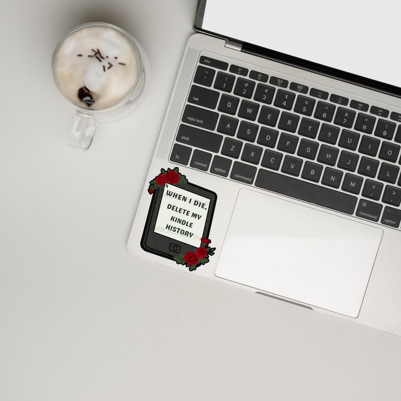 May include: A rectangular sticker featuring a black e-reader device surrounded by red roses and green leaves. The screen displays the black text: WHEN I DIE, DELETE MY KINDLE HISTORY. The sticker is placed on the palm rest area of a silver laptop.