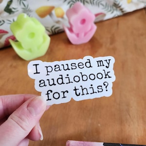 White sticker with black text that reads "I paused my audiobook for this? vinyl sticker held in hand, waterproof glossy finish.