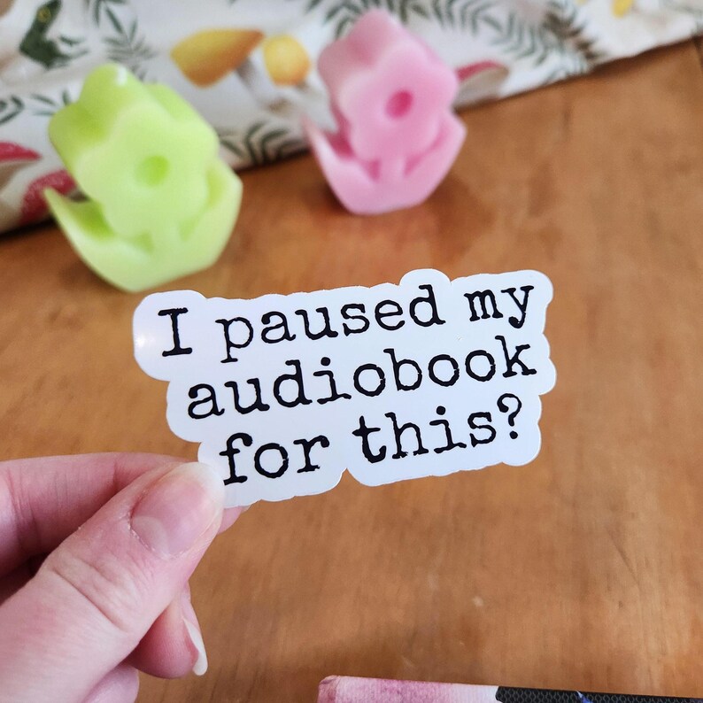 White sticker with black text that reads "I paused my audiobook for this? vinyl sticker held in hand, waterproof glossy finish.