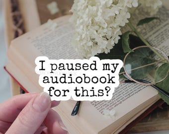 May include: A white die-cut sticker with black text that reads "I paused my audiobook for this?" positioned over an open book with cream-colored pages, a pair of thin-rimmed round glasses, and a cluster of white hydrangea flowers.