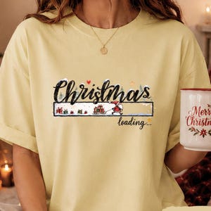 Puede incluir: Una camiseta de color amarillo pálido con un estampado gráfico que incluye el texto Christmas loading. El diseño muestra una barra de carga con un pequeño Santa Claus tirando de un trineo lleno de regalos, rodeado de árboles, un corazón rojo y estrellas.