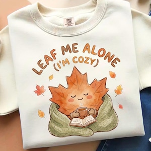 May include: A cream-coloured crewneck sweatshirt featuring a graphic of a smiling maple leaf character wrapped in a green blanket, holding a mug and reading an open book. The text above reads LEAF ME ALONE (I'M COZY) in brown letters.