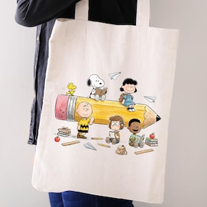 May include: Cream-colored tote bag featuring a yellow pencil design with cartoon characters. The pencil has a pink eraser and black tip. The characters include Snoopy, Charlie Brown, Lucy, and Woodstock. Paper airplanes and books are also shown.