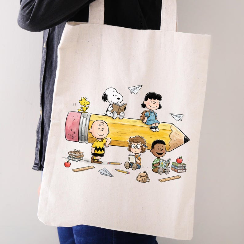 May include: Cream-colored tote bag featuring a yellow pencil design with cartoon characters. The pencil has a pink eraser and black tip. The characters include Snoopy, Charlie Brown, Lucy, and Woodstock. Paper airplanes and books are also shown.