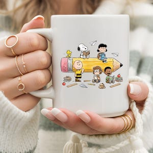 May include: White ceramic mug featuring a colorful illustration of Peanuts characters on a giant pencil. The design includes Snoopy, Charlie Brown, Lucy, and others, with books and paper airplanes. A fun, whimsical design for fans of the comic strip.