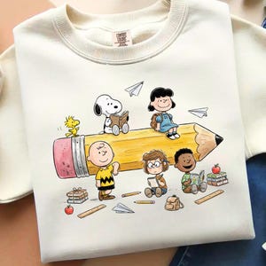 May include: Cream-colored sweatshirt featuring a colorful graphic of Peanuts characters. Snoopy, Woodstock, Charlie Brown, and others are depicted on a large pencil, with books, paper airplanes, and school supplies. The sweatshirt has a crew neck.