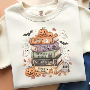 May include: A cream-coloured crewneck sweatshirt featuring a graphic print of a stack of five books titled Ghostly Tales, Spells & Potions, Haunted Houses, Witch's Brew, and Pumpkin Lore, surrounded by pumpkins, ghosts, bats, and spiderwebs.