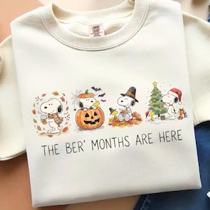 May include: A cream-colored crewneck sweatshirt featuring a graphic of Snoopy in four seasonal scenes, including autumn leaves, a jack-o'-lantern, a pilgrim hat, and a Christmas tree. The text below the illustrations reads, THE BER' MONTHS ARE HERE.