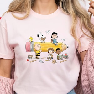 May include: Pink t-shirt featuring a colorful illustration of Peanuts characters. The design includes Snoopy, Charlie Brown, Lucy, and other characters sitting on a large yellow pencil, with books and paper airplanes scattered around.