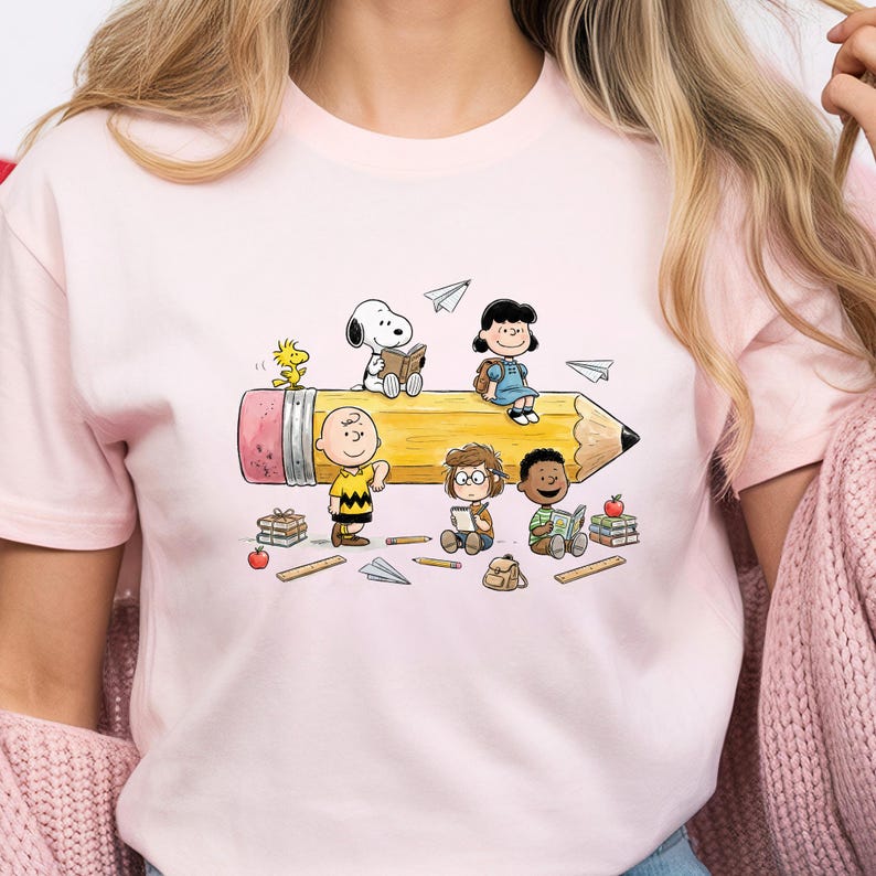 May include: Pink t-shirt featuring a colorful illustration of Peanuts characters. The design includes Snoopy, Charlie Brown, Lucy, and other characters sitting on a large yellow pencil, with books and paper airplanes scattered around.