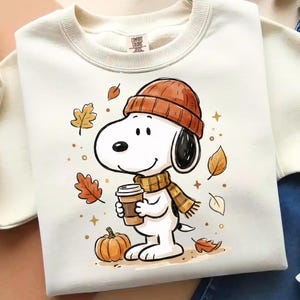 Op de afbeelding: Een crèmekleurig sweatshirt met ronde hals en een grafische illustratie van een witte tekenfilmhond met een oranje gebreide muts en een bijpassende sjaal. De hond houdt een bruine koffiebeker vast naast een oranje pompoen en herfstbladeren.