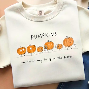 May include: A cream-coloured crewneck sweatshirt featuring a graphic illustration of six orange pumpkins with faces and limbs. The text reads PUMPKINS on their way to spice the lattes. The pumpkins are depicted walking, some carrying bags or briefcases.