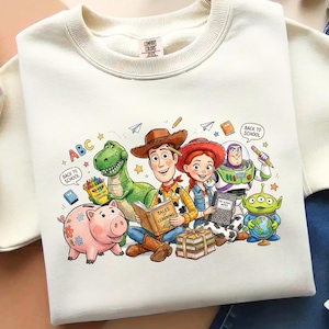 Puede incluir: Sudadera crema con una colorida ilustración de los personajes de Toy Story. El diseño incluye a Woody, Jessie, Buzz Lightyear, Rex y otros personajes con libros y elementos escolares. También se incluye el texto "ABC" y "Back to School".