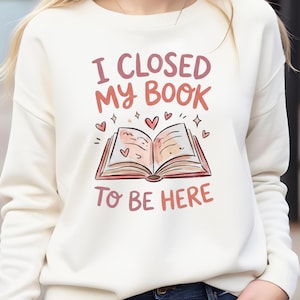Peut inclure: Sweat-shirt crème avec l'inscription "I CLOSED MY BOOK TO BE HERE" en rose et rouge. Le motif représente un livre ouvert avec des cœurs et des étoiles. Un haut confortable pour les amateurs de lecture.