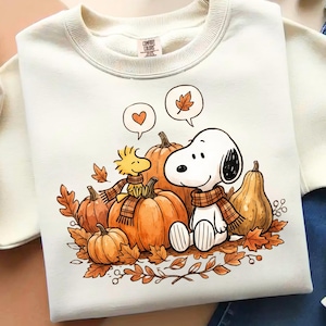 May include: A cream-colored crewneck sweatshirt featuring a graphic print of Snoopy and Woodstock sitting among orange pumpkins and brown autumn leaves. Both characters wear plaid scarves, and speech bubbles show a heart and a leaf.