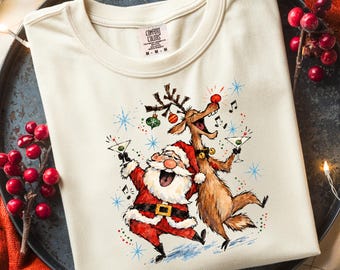 May include: A cream-colored short-sleeve t-shirt featuring a graphic print of Santa Claus and a reindeer dancing while holding martini glasses. The design includes musical notes, colorful ornaments, and blue starbursts. The shirt has a Comfort Colors label.
