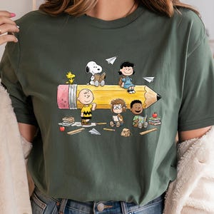 May include: Dark green t-shirt featuring a cartoon illustration of the Peanuts gang riding a giant pencil. Snoopy, Charlie Brown, Lucy, and other characters are depicted with books and paper airplanes. The pencil has a pink eraser and a sharpened graphite tip.