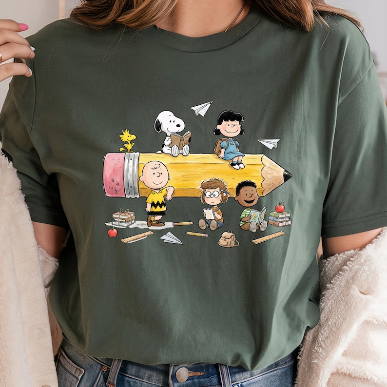 May include: Dark green t-shirt featuring a cartoon illustration of the Peanuts gang riding a giant pencil. Snoopy, Charlie Brown, Lucy, and other characters are depicted with books and paper airplanes. The pencil has a pink eraser and a sharpened graphite tip.