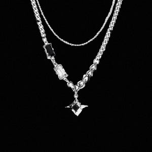 May include: A layered silver-tone necklace featuring a delicate chain and a thicker curb chain. The thicker chain is adorned with two rectangular crystals, one black and one clear, and a central four-pointed star pendant with a black stone.