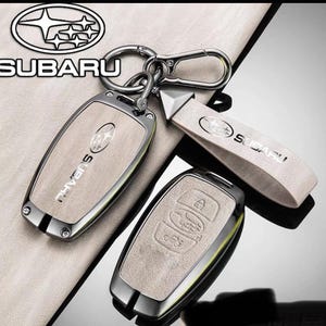 Peut inclure: Un ensemble de coques de clé Subaru beige avec un cadre métallique et une lanière assortie. La coque affiche le logo et le texte Subaru, avec une pièce secondaire montrant des icônes de boutons en relief pour verrouiller et déverrouiller.