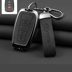 May include: A black car key fob with a metallic silver frame and a matching black leather keychain strap featuring the embossed Toyota logo. The fob has four buttons with icons for locking, unlocking, trunk release, and a panic alarm, plus a metal clip.