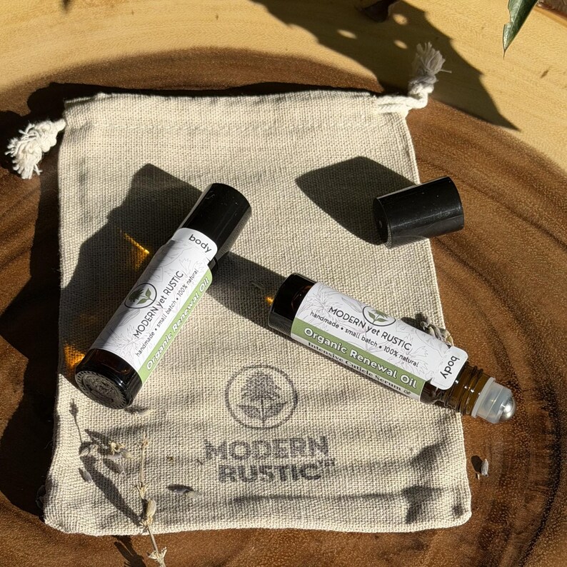 Puede incluir: Dos botellas roll-on de vidrio de "Organic Renewal Oil" con tapas y etiquetas negras, junto a una bolsa beige con cord&oacute;n. Las etiquetas dicen "MODERN yet RUSTIC" y "handmade small batch 100% natural". La bolsa tiene un logotipo y las palabras "MODERN RUSTIC".