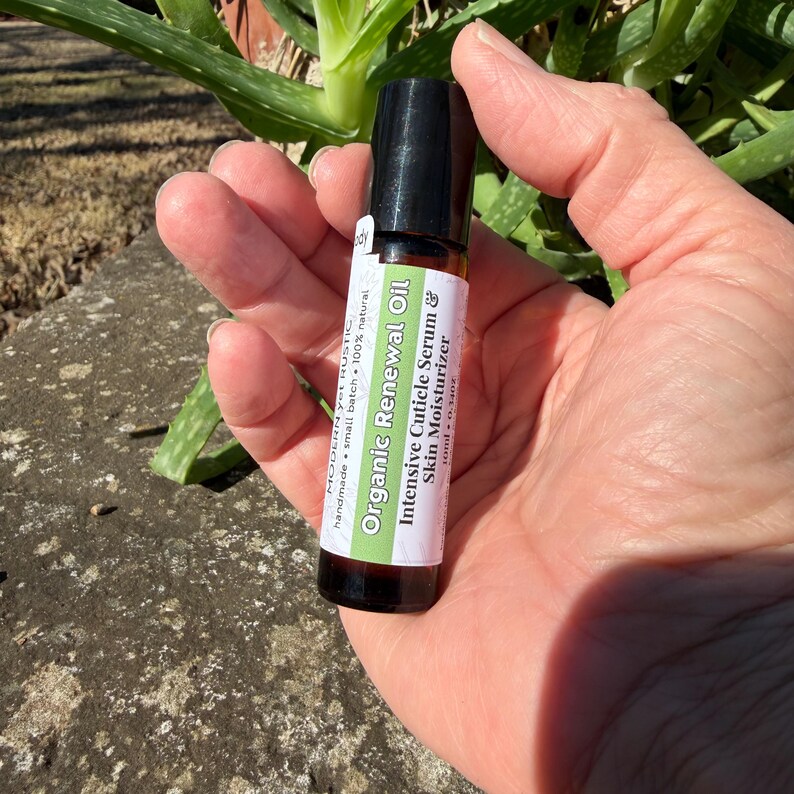 Puede incluir: Un peque&ntilde;o frasco de vidrio transparente con tapa negra se sostiene en una mano. El frasco tiene una etiqueta blanca con texto verde y negro que dice "Organic Renewal Oil" y "Intensive Cuticle Serum & Skin Moisturizer".