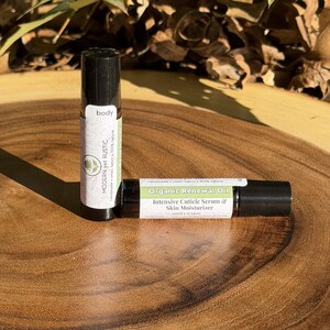 Puede incluir: Dos botellas roll-on de aceite corporal sobre una superficie de madera. Una botella est&aacute; en posici&oacute;n vertical, etiquetada como "body" y "MODERN yet RUSTIC". La otra botella est&aacute; en posici&oacute;n horizontal, etiquetada como "Organic Renewal Oil" y "Intensive Cuticle Serum & Skin Moisturizer".