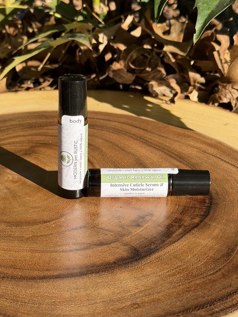 Puede incluir: Dos botellas roll-on de aceite corporal sobre una superficie de madera. Una botella est&aacute; en posici&oacute;n vertical, etiquetada como "body" y "MODERN yet RUSTIC". La otra botella est&aacute; en posici&oacute;n horizontal, etiquetada como "Organic Renewal Oil" y "Intensive Cuticle Serum & Skin Moisturizer".