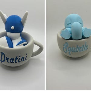 May include: Two ceramic teacups, each with a different Pok&eacute;mon figure inside. One cup has a blue and white Dratini, the other a light blue Squirtle. The names of the Pok&eacute;mon are written on the cups in blue.