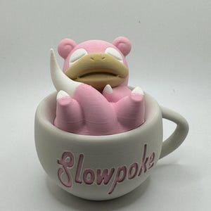May include: A pink Slowpoke figure rests inside a gray teacup with the word "Slowpoke" in pink script. The figure has a tan snout and white accents. The teacup has a handle.