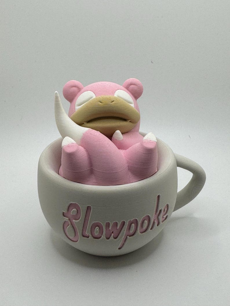 May include: A pink Slowpoke figure rests inside a gray teacup with the word "Slowpoke" in pink script. The figure has a tan snout and white accents. The teacup has a handle.