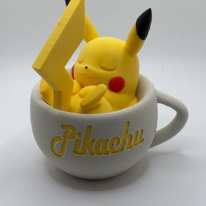May include: A yellow Pikachu figure with red cheeks and black-tipped ears rests inside a white teacup. A yellow lightning bolt is positioned behind the character. The word "Pikachu" is written in yellow script on the cup.