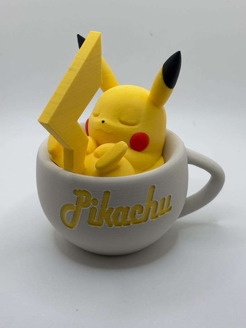May include: A yellow Pikachu figure with red cheeks and black-tipped ears rests inside a white teacup. A yellow lightning bolt is positioned behind the character. The word "Pikachu" is written in yellow script on the cup.