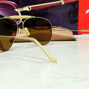 May include: Gold-tone aviator sunglasses featuring brown tinted lenses, a thin metal frame, and a distinct brow bar. The temples are slender with matching gold-tone tips, and the nose pads are light-colored. The design includes a classic teardrop shape.