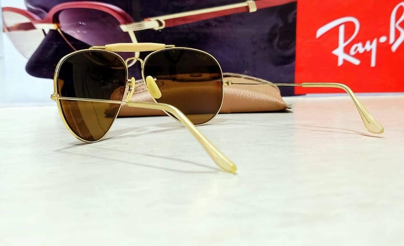 May include: Gold-tone aviator sunglasses featuring brown tinted lenses, a thin metal frame, and a distinct brow bar. The temples are slender with matching gold-tone tips, and the nose pads are light-colored. The design includes a classic teardrop shape.